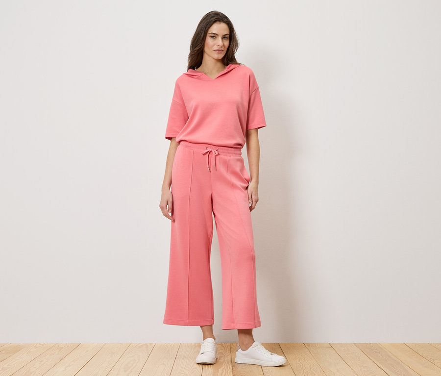 Egy nő áll a fal előtt, korall színű, kétrészes ruhát visel, amely egy rövid ujjú kapucnis felsőből és egy culottes nadrágból áll, valamint fehér tornacipőt.