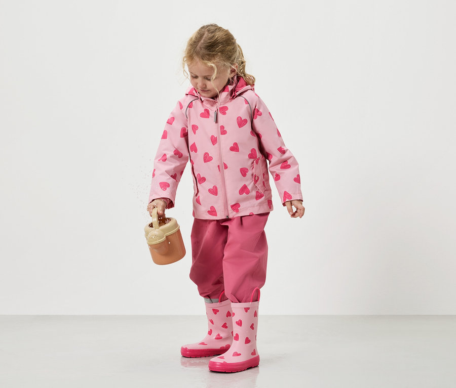 Egy kislány homokot önt ki egy játékvödörből, rózsaszín, szívecskés mintás kabátban, hozzáillő nadrágban és gumicsizmában.