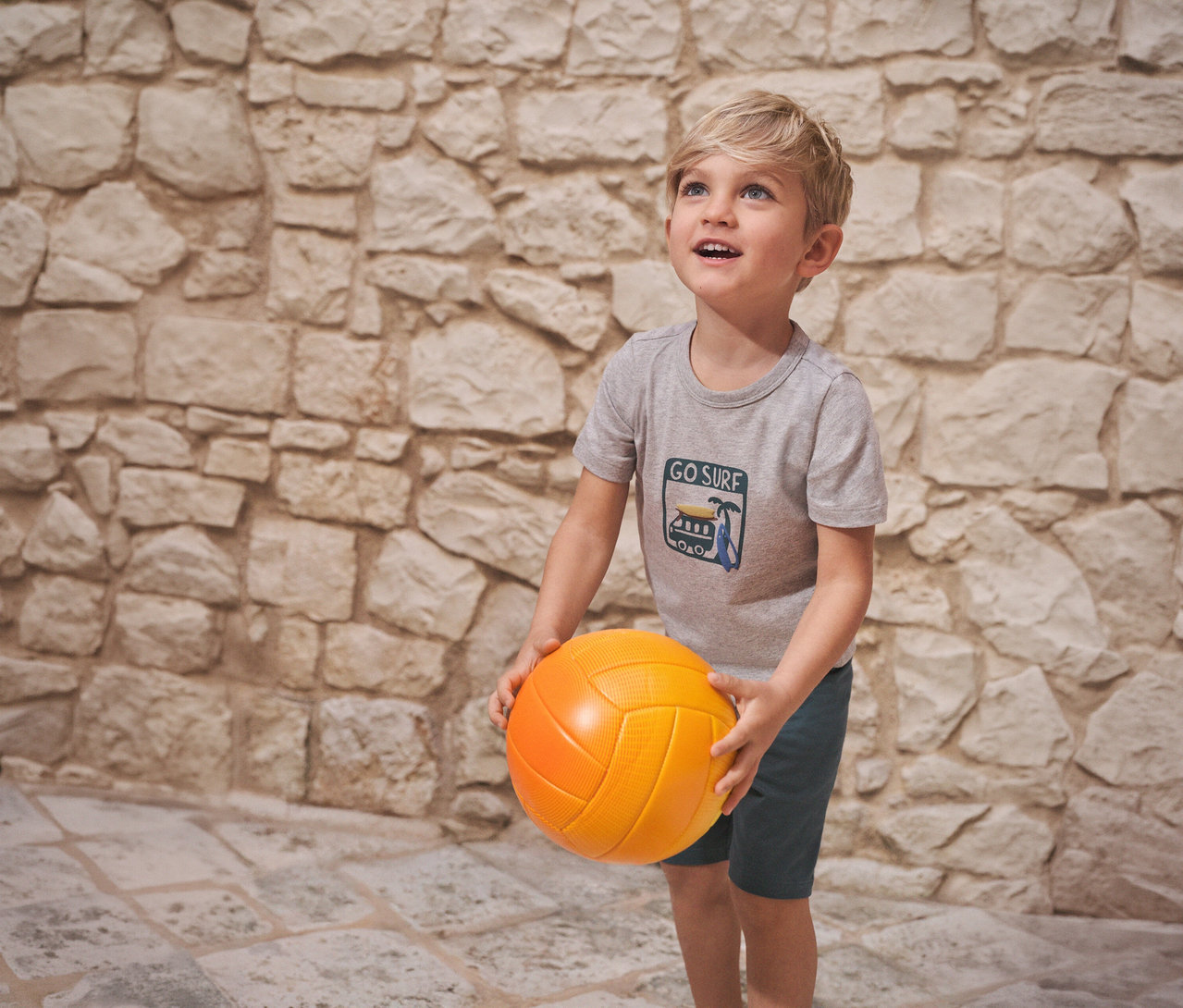 Fiú narancssárga labdát tart, és SCHIESSER rövidnadrágos gyerekpizsamát visel egy kőfal előtt.