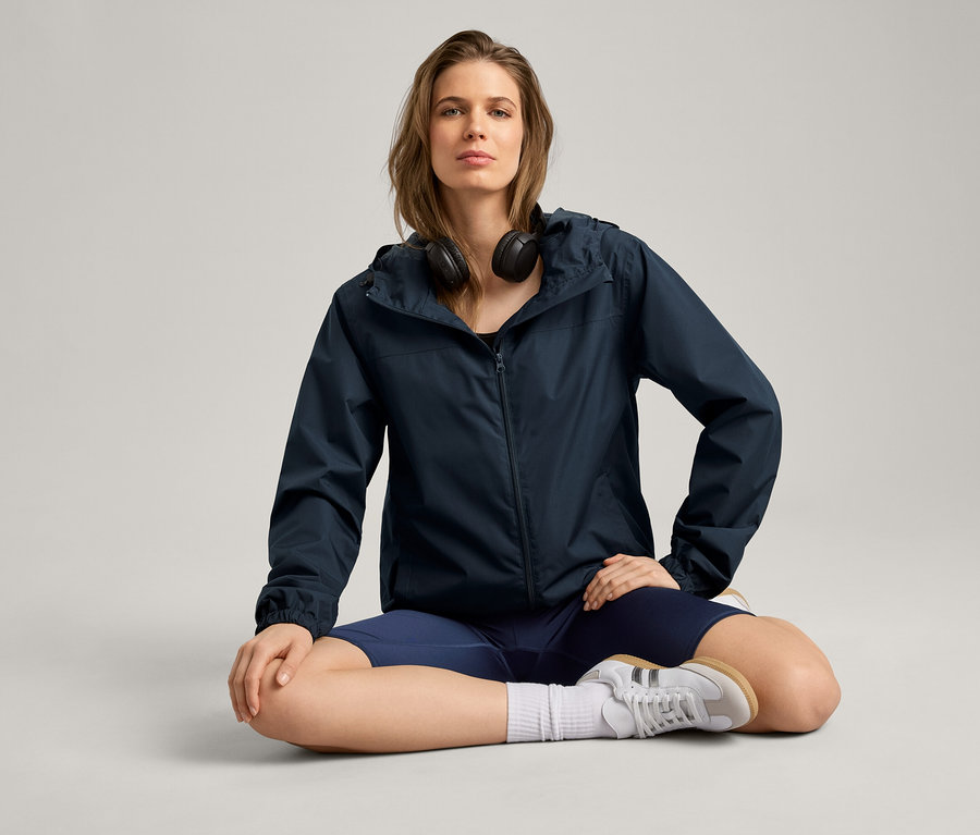 Nő ül törökülésben, esőkabátot, fejhallgatót, rövidnadrágot és tornacipőt visel.