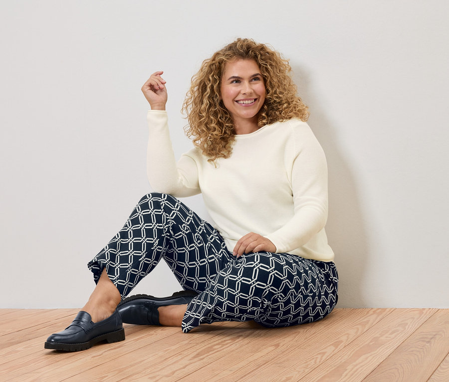 Egy göndör hajú nő ül egy fa padlón, krémszínű pulóvert és kék, geometriai mintás nadrágot visel.