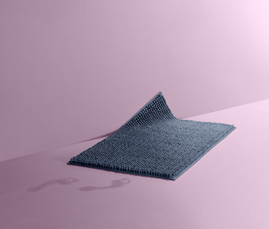 Egy kék kádkilépő, kb. 50 x 80 cm-es méretben, rózsaszín háttér előtt. A padlón nedves lábnyomok láthatók.
