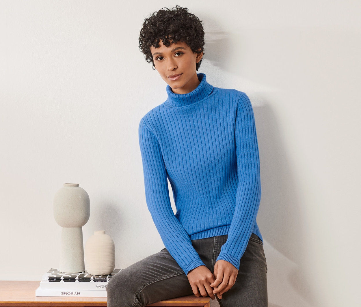 Nő ül egy fa komódon, kék garbónyakú pulóvert visel.