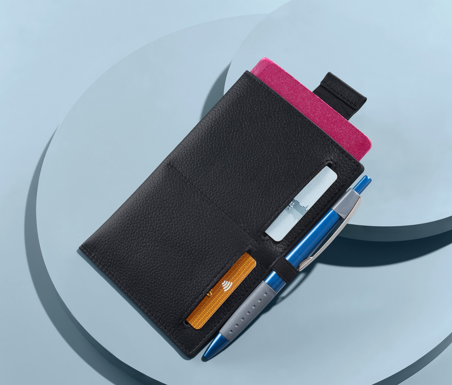 Fekete bőr útlevéltartó tok útlevéllel, kártyákkal és tollal kék háttér előtt.