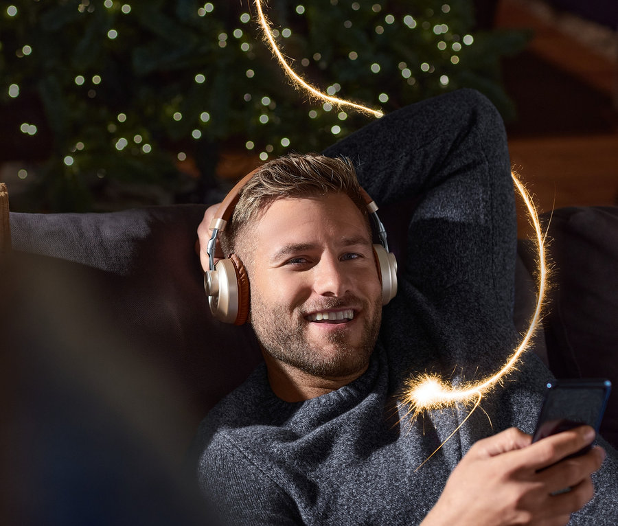 Mosolygó férfi fekszik a kanapén Bluetooth fejhallgatóval a fülén, kezében telefon. A háttérben karácsonyfa.
