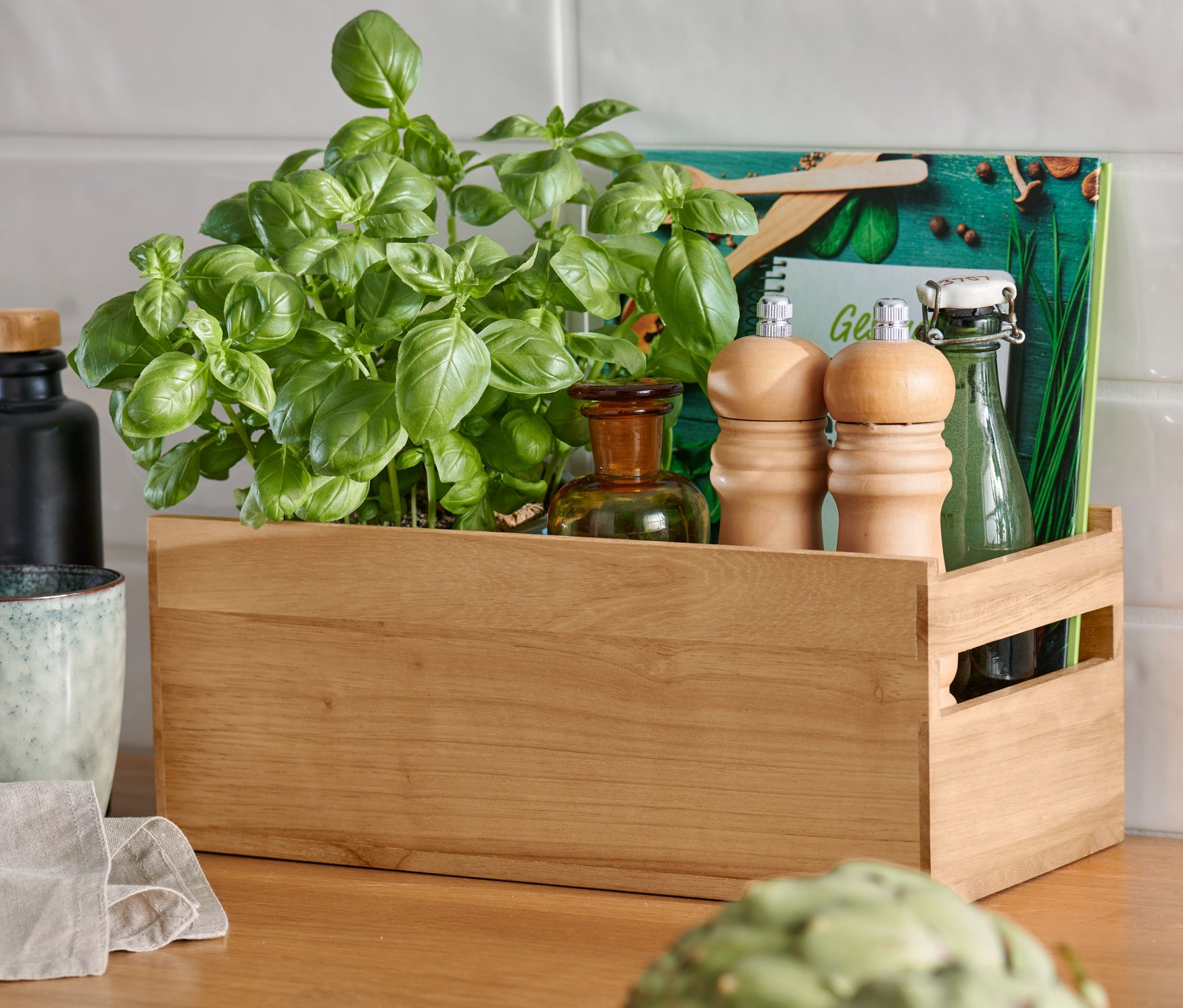 Egy fából készült fűszertartó doboz bazsalikommal, fűszermalmok és üvegpalackok állnak egy fa konyhapulton, előtérben egy articsóka.