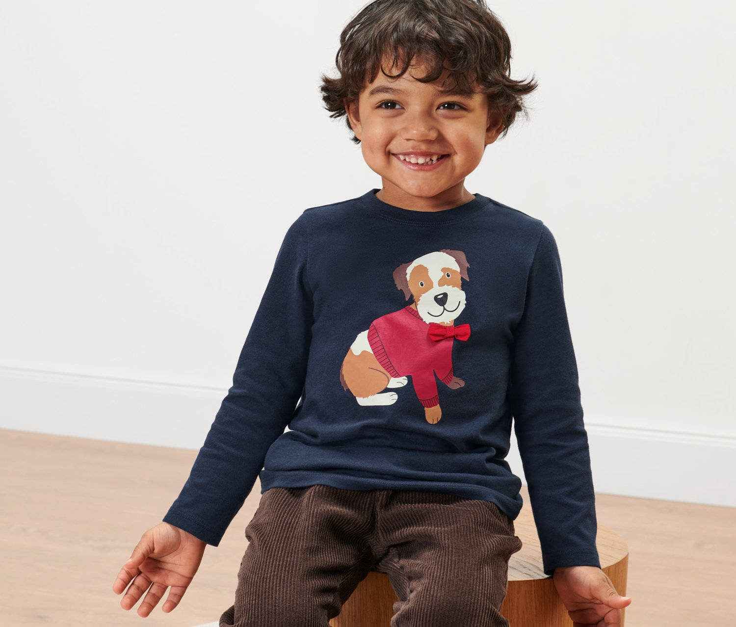 Egy mosolygó kisfiú ül, és kutyás kisgyerek hosszú ujjú felsőt visel.