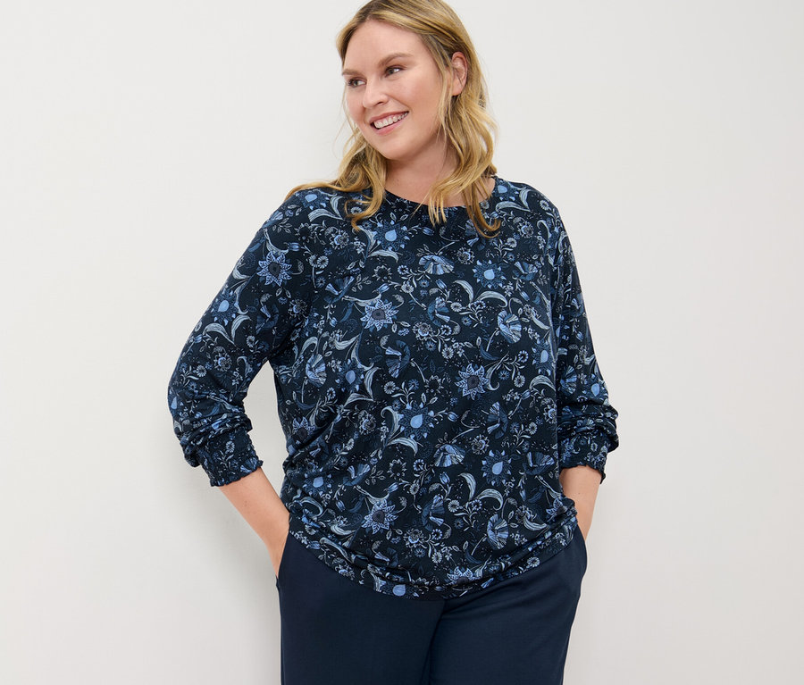 Nő virágos hosszú ujjú pólóblúzban és sötétkék jersey nadrágban pózol.