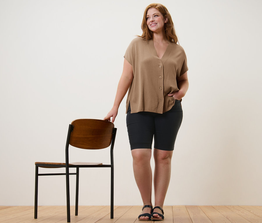 Egy nő egy szék mellett áll, bézs rövid ujjú kardigánt és fekete női bengalin bermudanadrágot visel.