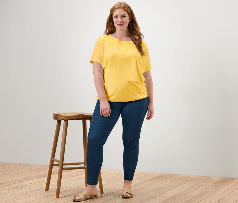 Nő pózol egy szék mellett, sárga, hímzett pólóblúzt és női jeggingst visel.