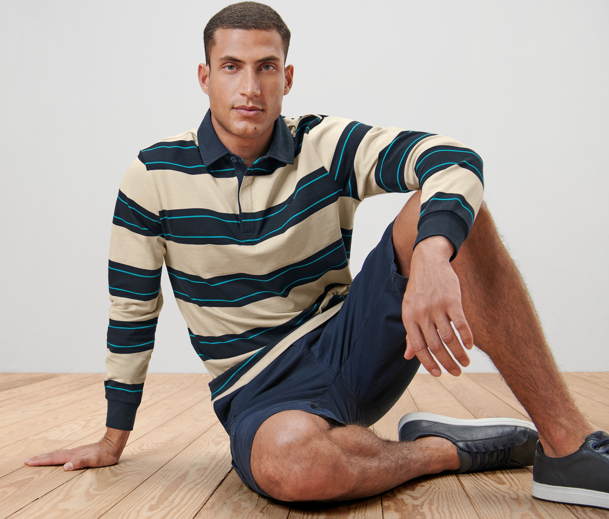 Férfi ül a fapadlón, Férfi hosszú ujjú pólóing, csíkos, sötétkék/bézs rövidnadrágot és tornacipőt visel.