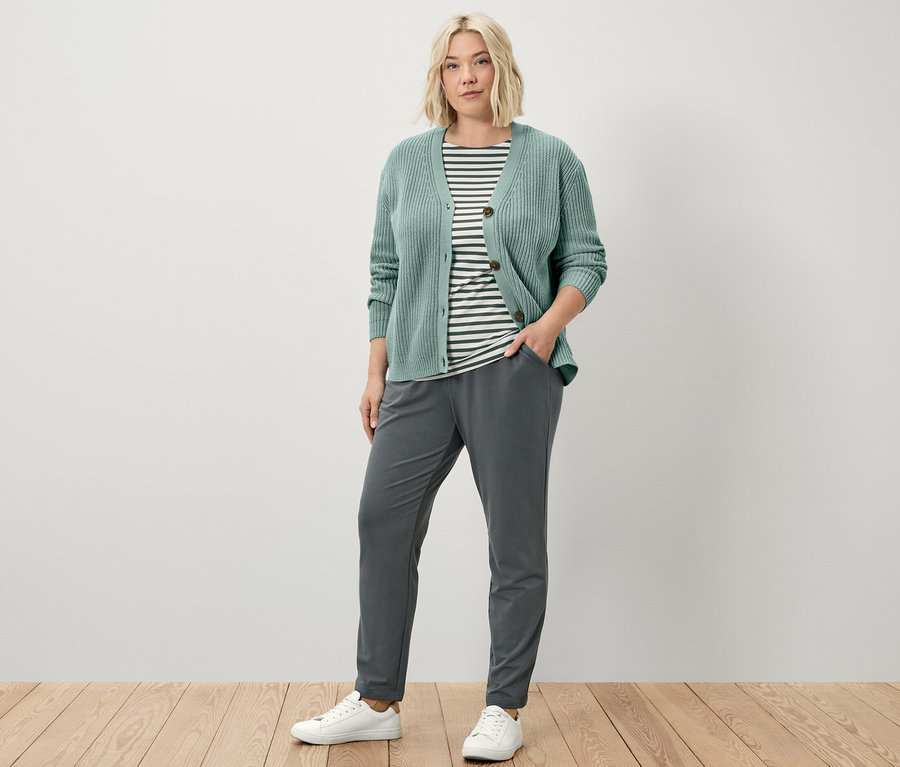 Egy nő áll, és szabadidőnadrágot, laza kötésű, zsályazöld kardigánt és zöld csíkos, félhosszú ujjú felsőt visel.