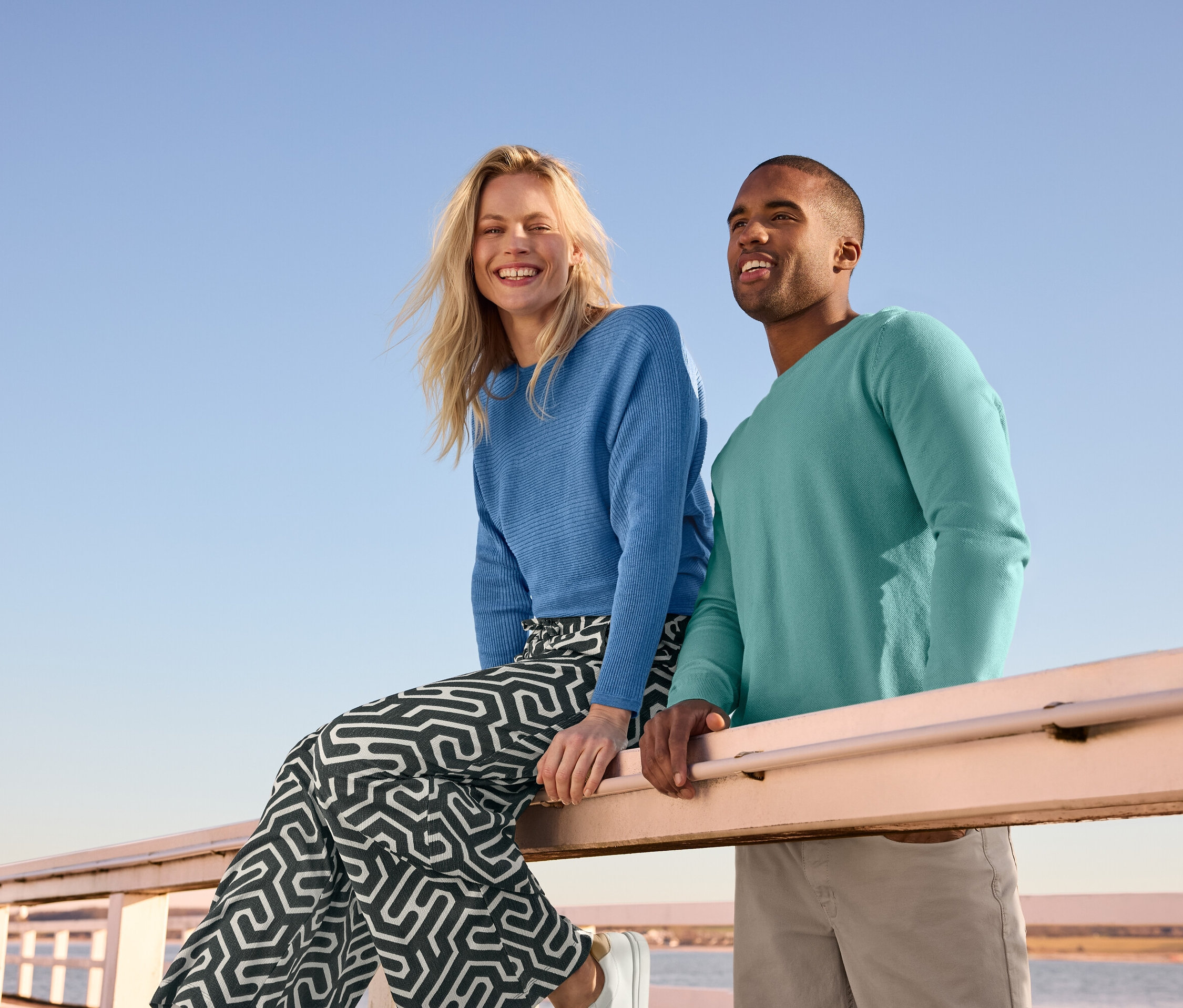 Egy nő egy korláton ül, kék, finom kötésű pulóvert és nyomott mintás, gyűrött hatású belebújós nadrágot visel. Mellette egy férfi áll kötött pulóverben és Férfi nadrágban, bézs színben.