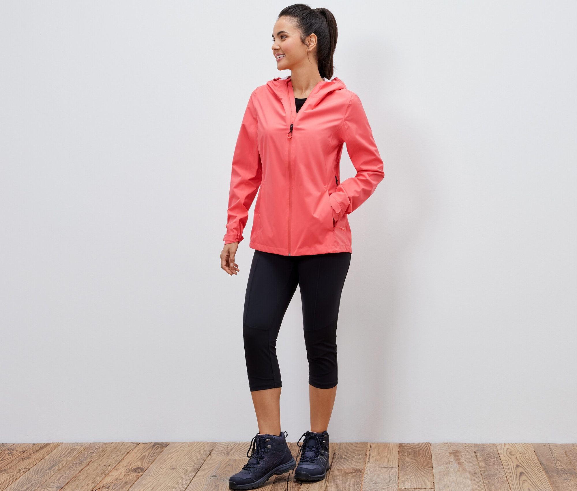 Egy mosolygó nő korall színű 2,5 rétegű szabadidős kabátban, fekete háromnegyedes leggingsben és túrabakancsban pózol.