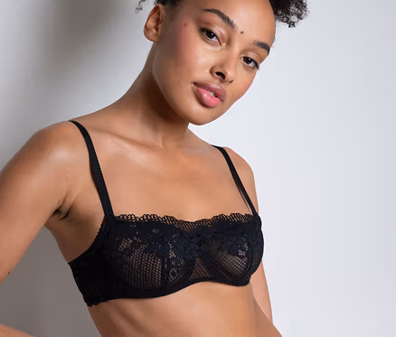 Egy nő feltűzött göndör hajjal fekete Passionata bandeau csipke melltartó »Nina«-t és hozzá illő alsóneműt visel, egyik kezét a csípőjén tartja.