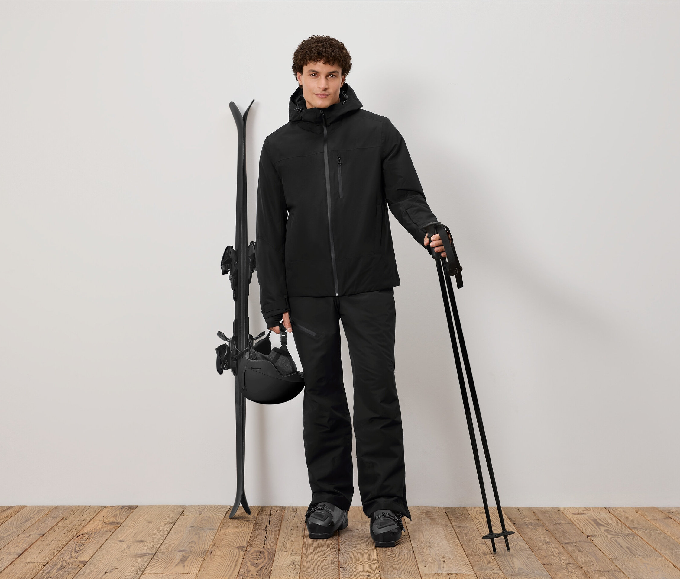 Férfi fekete síruhában botokat tart, mellette síléc és sisak.