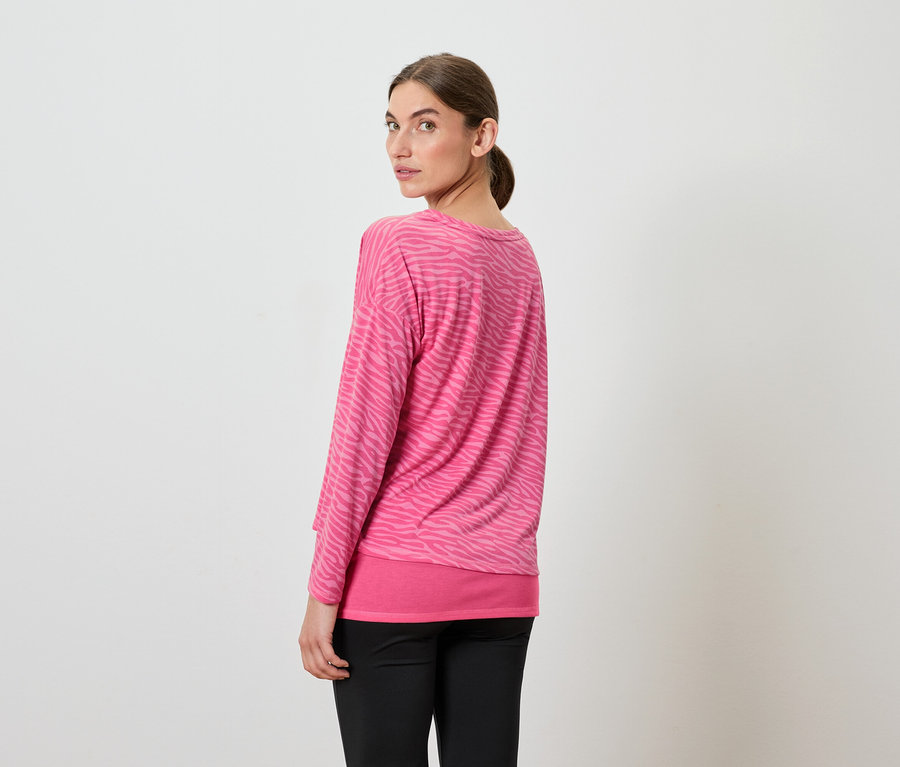 Egy nő visszanéz a válla fölött, rózsaszín, hosszú ujjú felsőt és fekete nadrágot visel.