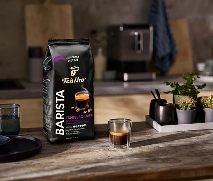 Egy csomag Tchibo Barista Espresso Dark kávébab áll egy eszpresszós pohár mellett egy fa asztalon.