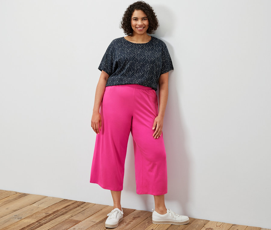 Egy nő áll, sötétkék, rövid ujjú ejtett vállú pólóblúzt és rózsaszín bő szárú jerseynadrágot visel.