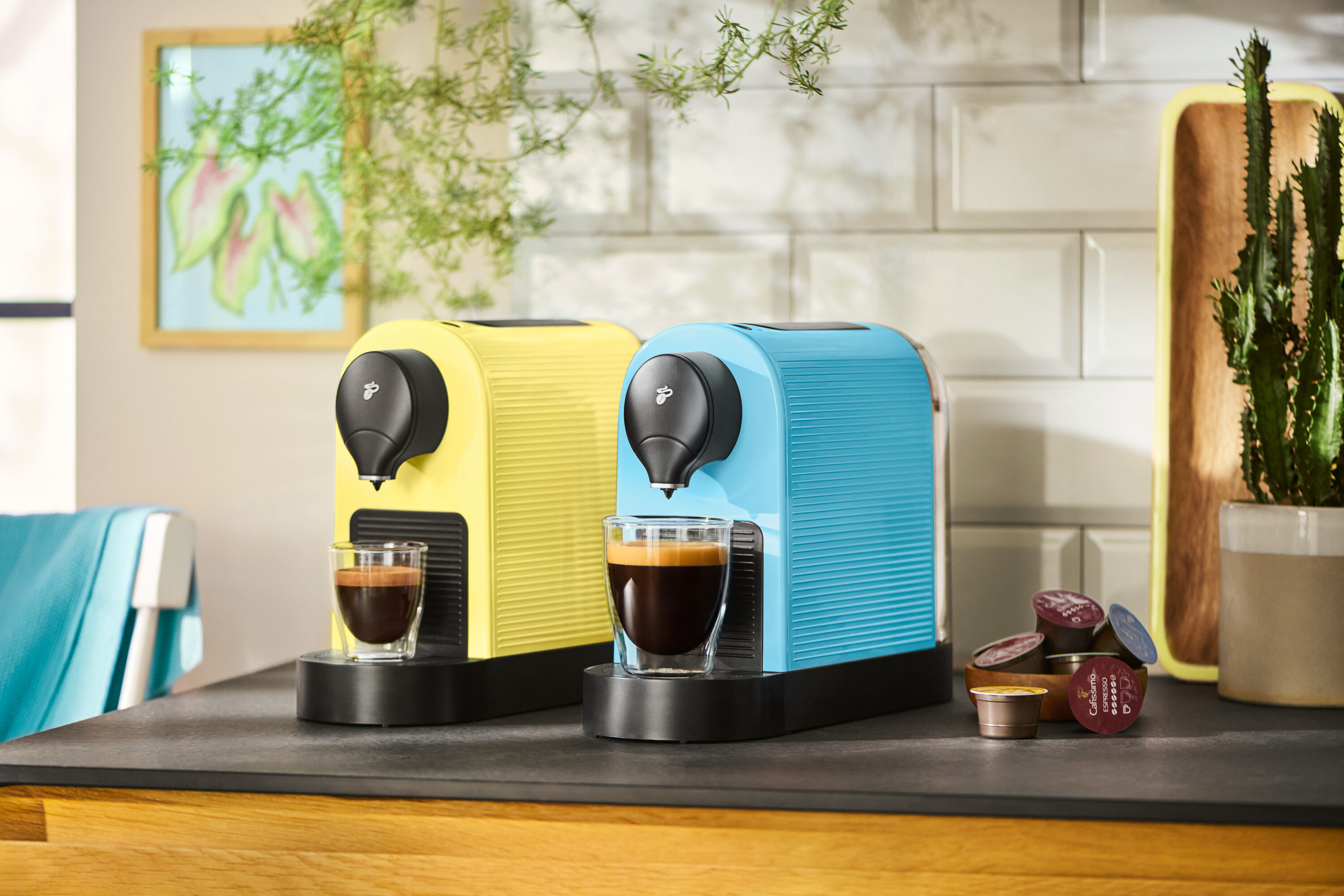 Két kávéfőző sárga és kék színben áll a konyhapulton két csésze kávéval és kávékapszulákkal.