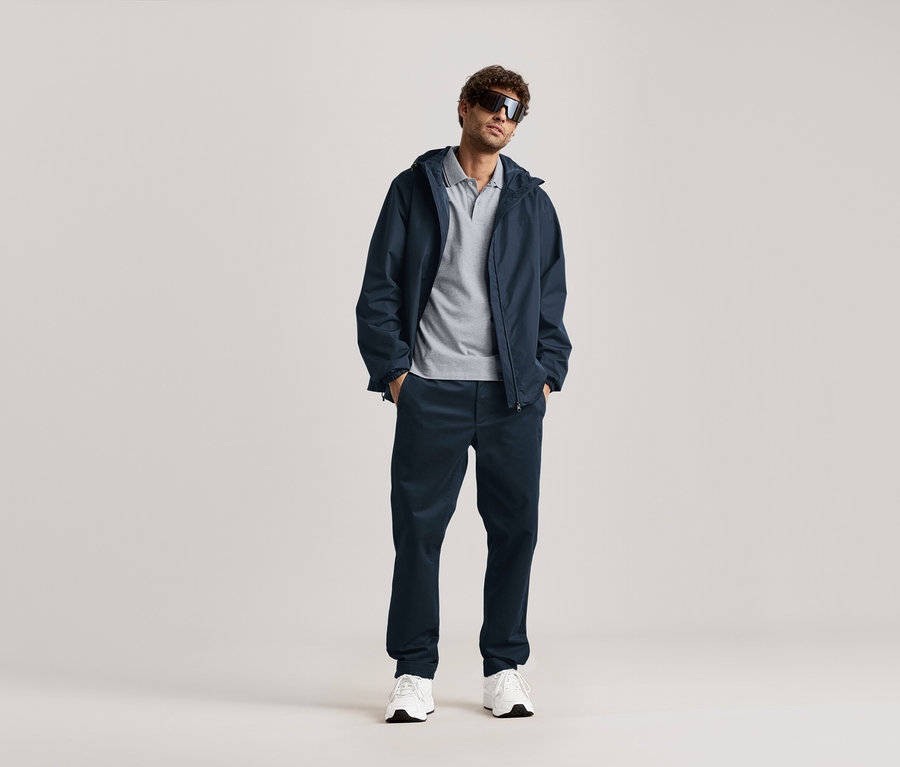 Egy férfi áll napszemüvegben, sötétkék kabátban és sötétkék chino nadrágban.