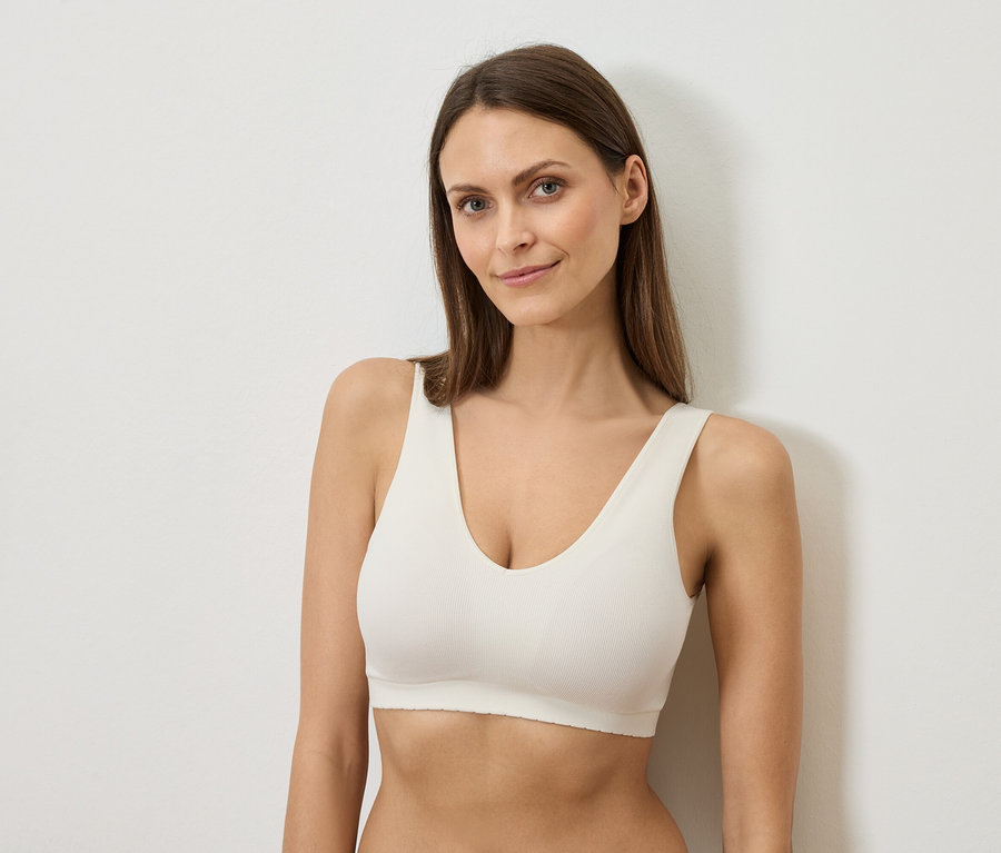 Hosszú barna hajú nő fehér melltartót visel és egy fehér falnak támaszkodik.