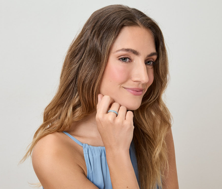 Hullámos hajú, kék felsőt viselő nő az ujját érinti, amelyen egy gyűrű van, világos háttér előtt.