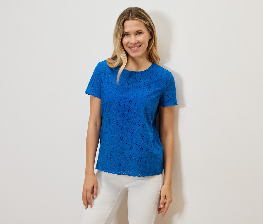 Egy mosolygó nő kék, rövid ujjú, lyukacsos mintázatú pólót és fehér nadrágot visel fehér háttér előtt.