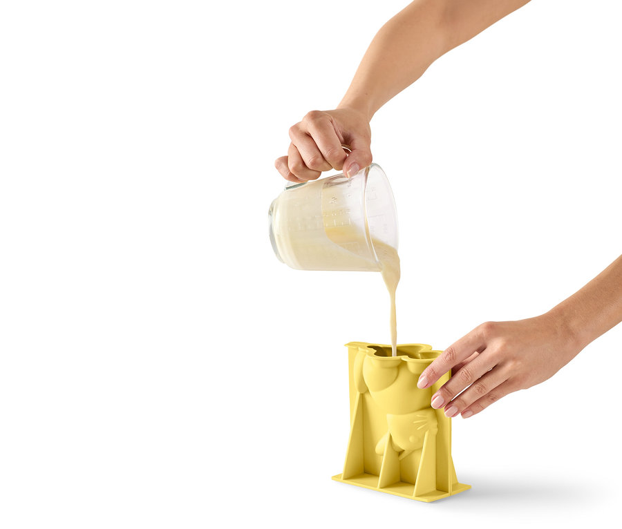 Egy személy tésztát önt egy mérőpohárból egy sárga, macska alakú szilikonformába.