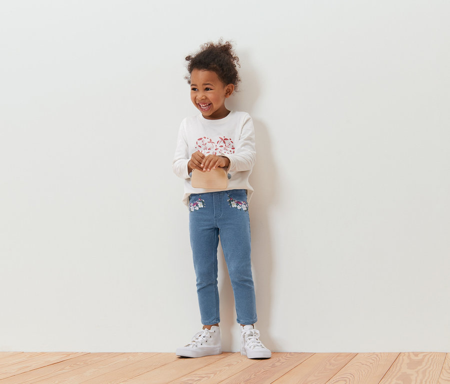 Mosolygó, göndör hajú kislány bézs színű kézitáskával, hosszú ujjú felsővel és virágmintás kék farmerrel, fehér fal előtt állva.