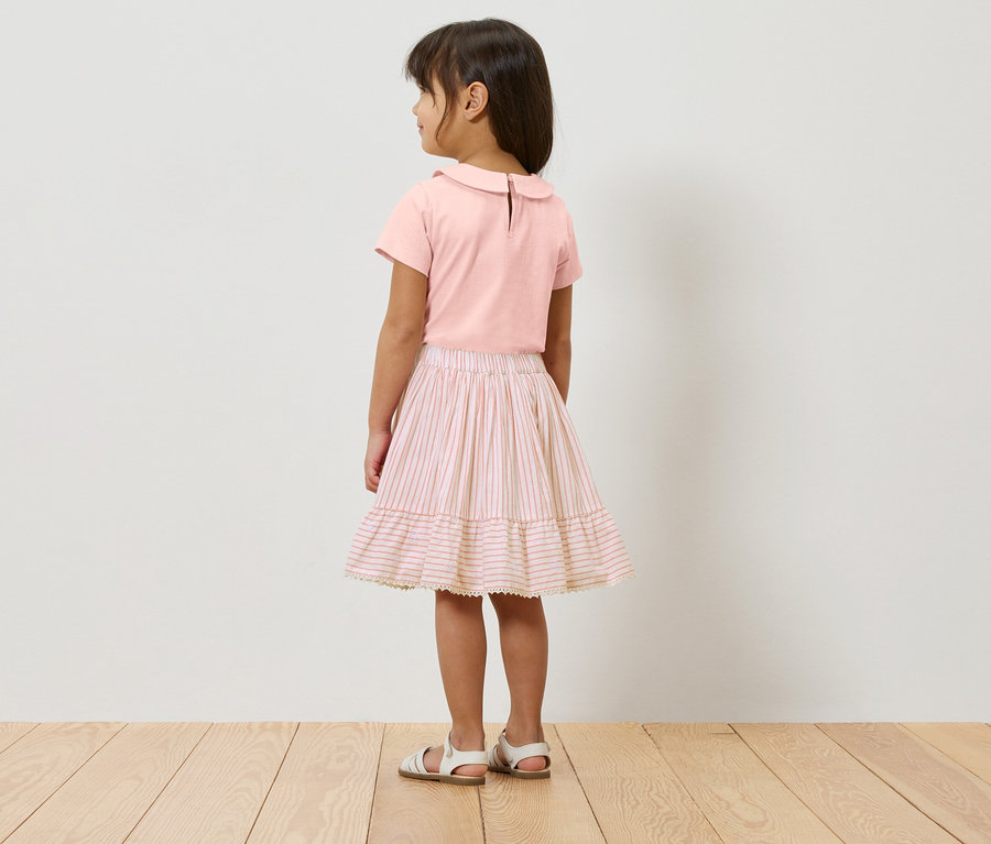Egy lány áll háttal a kamerának, rózsaszín felsőt, rózsaszín-fehér csíkos szoknyát és fehér szandált visel.