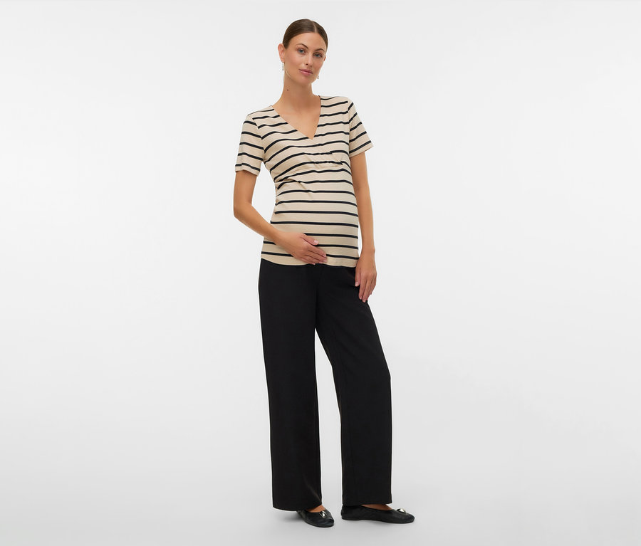 Egy nő a hasán tartja a kezét, fekete csíkos MAMALICIOUS »MLLANE TESS« kismama pólót, fekete nadrágot és balerinacipőt visel.