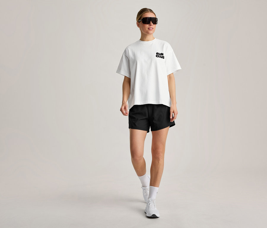 Egy nő sétál, fehér sportfelsőt visel „RUN CLUB” felirattal, fekete rövidnadrágot, fehér zoknit, fehér edzőcipőt és fekete napszemüveget.