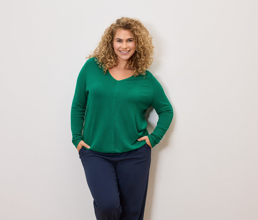 Göndör hajú nő zöld pulóvert és sötétkék nadrágot visel, kezei a zsebében.