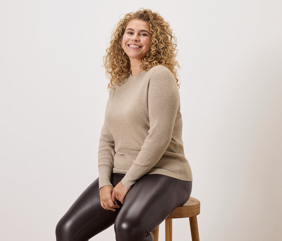 Egy göndör hajú nő ül egy fa zsámolyon, bézs pulóvert és sötét leggingset visel.