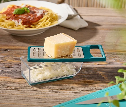 Egy fa asztalon egy tányér spagetti, egy sajtreszelő sajttal, egy vágódeszka bazsalikommal és cukkínivel.
