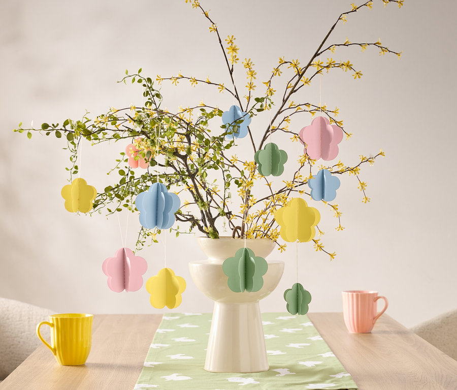 Virágdíszek lógnak egy ágon egy vázában az asztalon, egy sárga és egy rózsaszín bögre mellett.