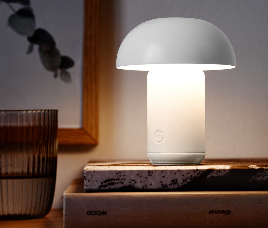 Egy fehér LED-es akkus asztali lámpa áll könyveken egy pohár víz mellett.