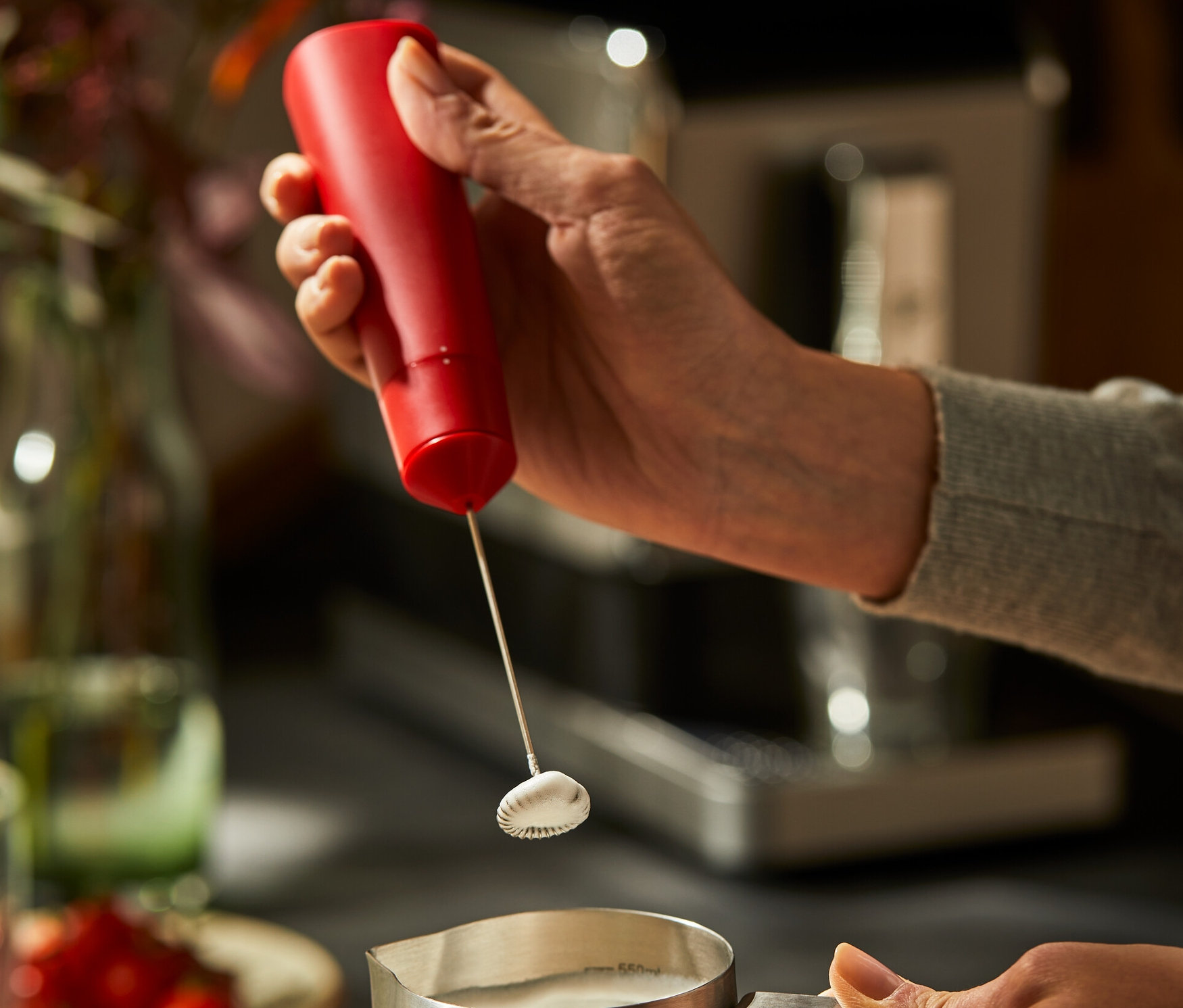 Egy nő egy piros elemes tejhabosítóval habosítja a tejet egy rozsdamentes acél kancsóban.