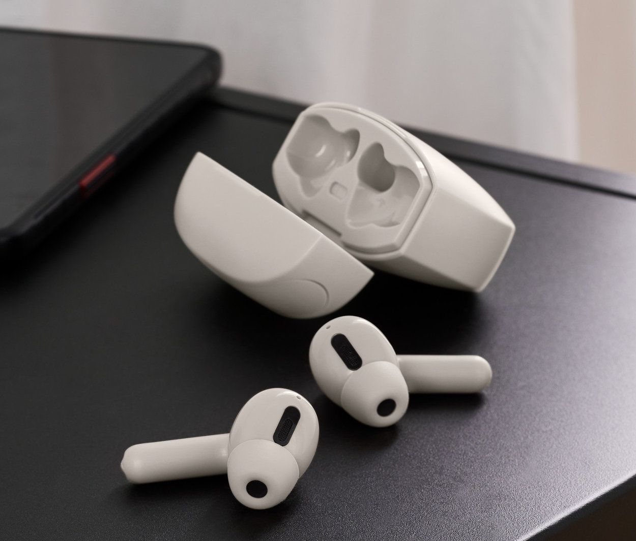 A fekete asztallapon vezeték nélküli fülhallgató, világosszürke színben, egy telefon és egy váza virágokkal.