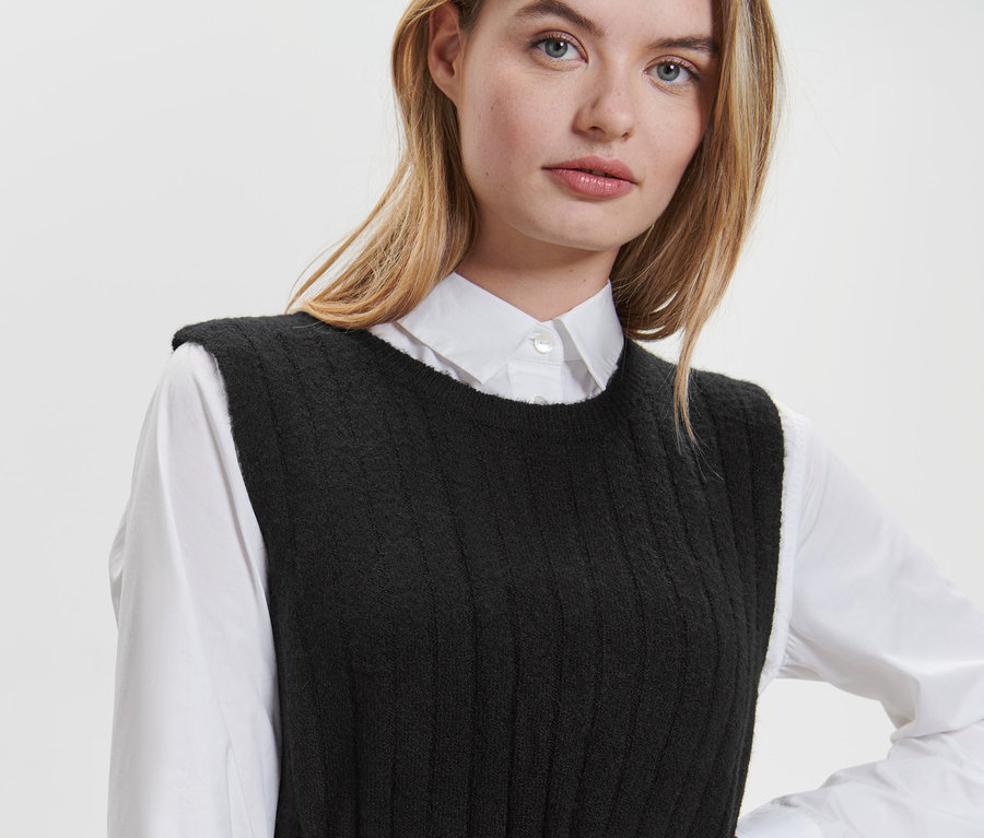 Egy szőke nő fekete, bordás kötésű ruhát visel egy fehér ing felett.