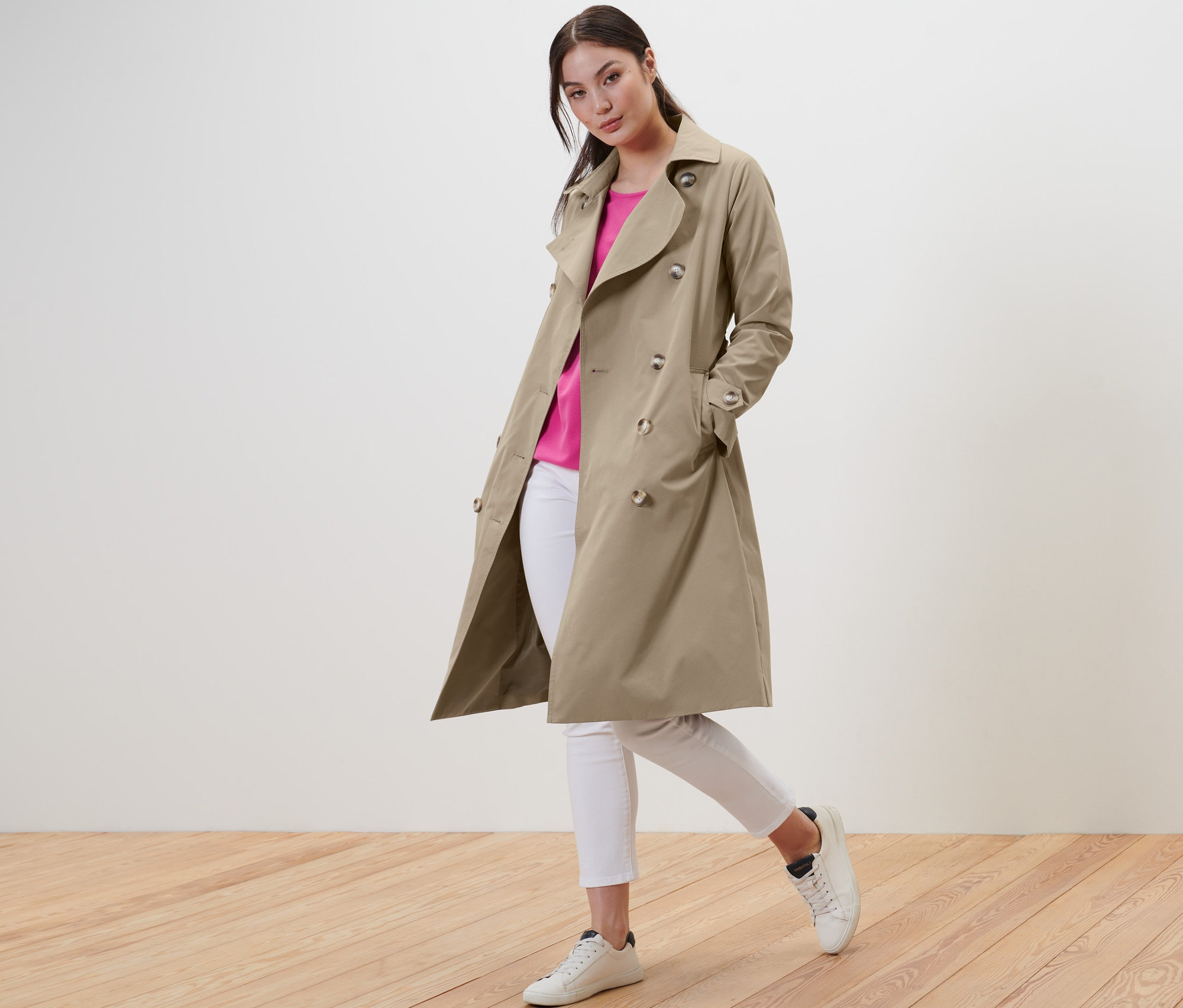 Nő pózol bézs ballonkabátban, fehér farmerban és tornacipőben.