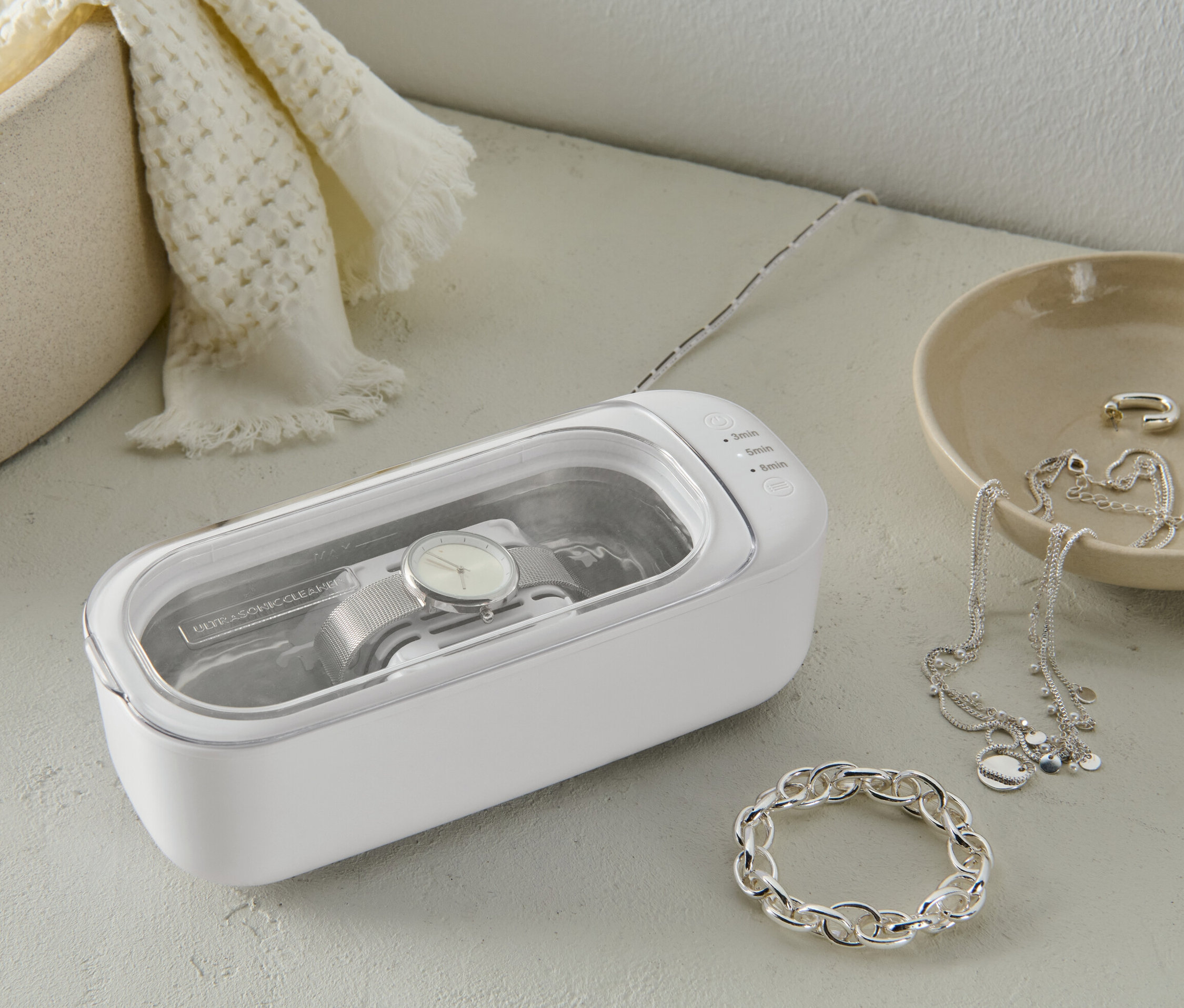 Egy óra tisztítása egy fehér ultrahangos tisztítóeszközben. Ékszerek vannak mellette.