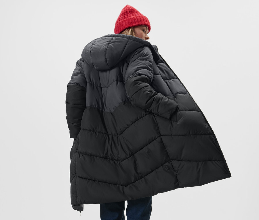 Nő visel fekete/szürke, steppelt kabátot kapucnival és piros sapkát.