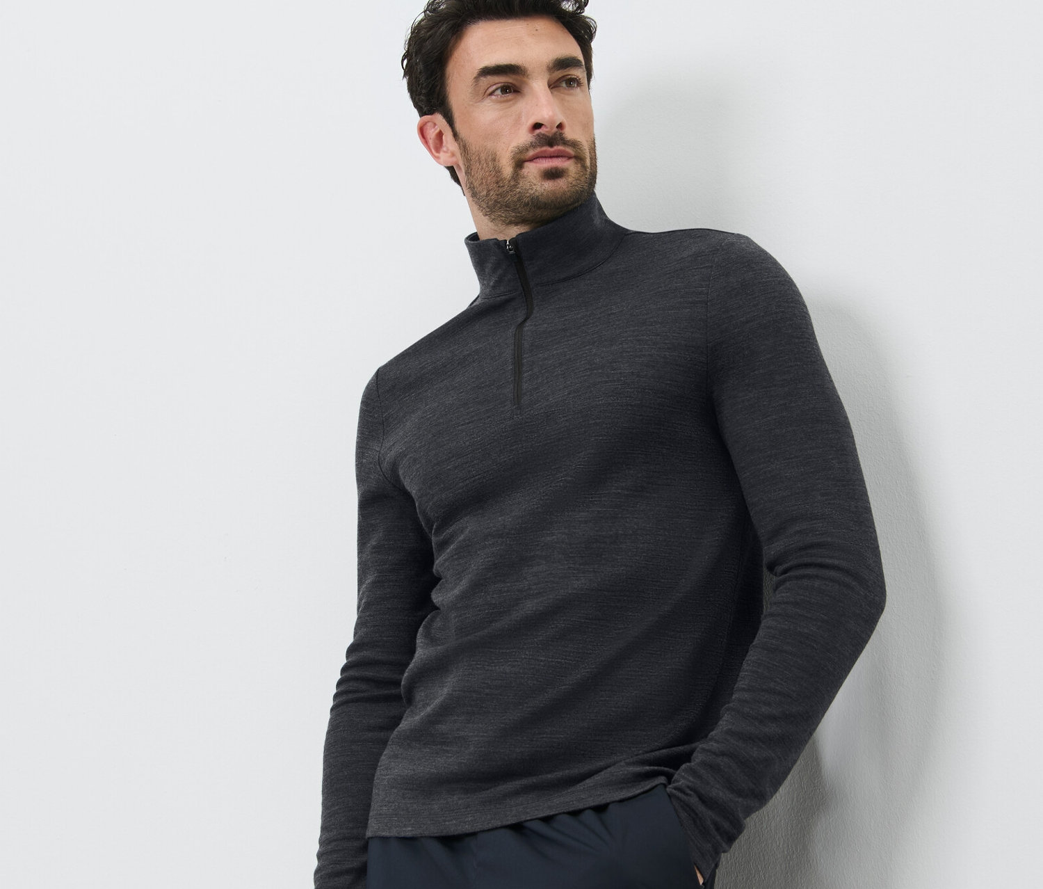 Férfi pózol fehér háttér előtt, RWS minősített gyapjúval készült multifunkcionális felsőt és sötét nadrágot visel.