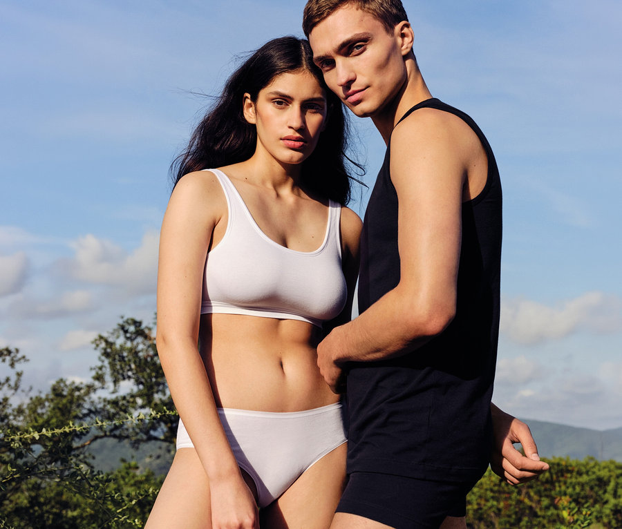 Férfi és nő áll a szabadban, a kamerába nézve. A nő fehér sportmelltartót és fehér SCHIESSER hipster alsót visel. A férfi fekete atlétát és fekete boxeralsót visel.