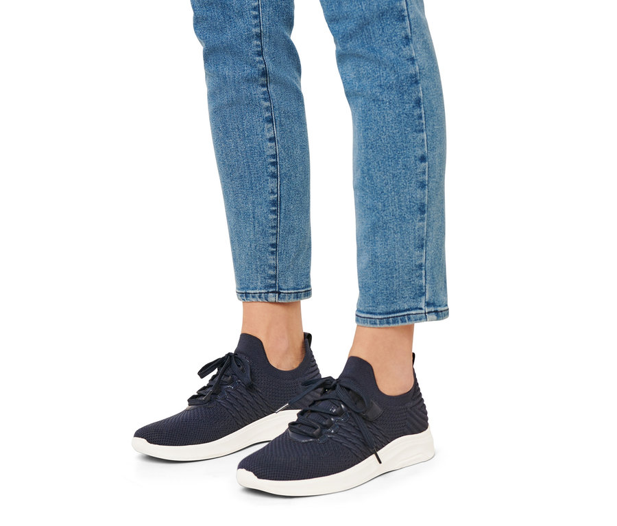 Egy személy lábai farmerban és kék, fehér talpú női sneaker cipőben.