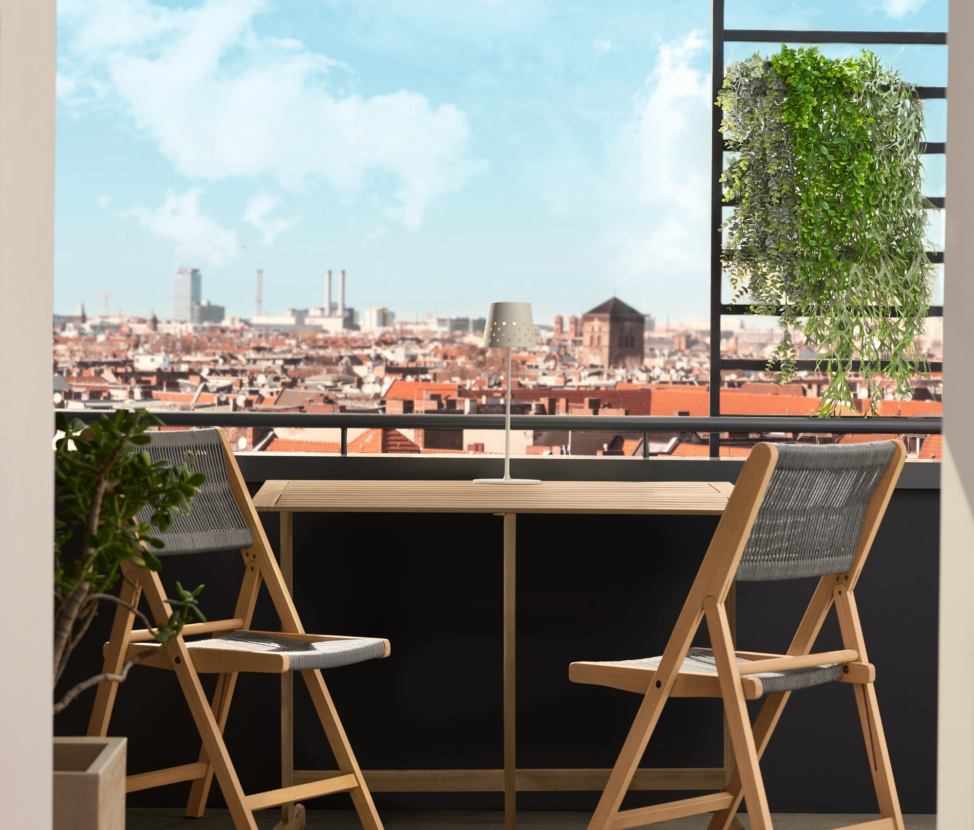 Összecsukható Lenja balkon asztal két Lenja textil hálós székkel és egy fém LED-es akkus asztali lámpával egy erkélyen. Egy fél napernyő árnyékot ad.