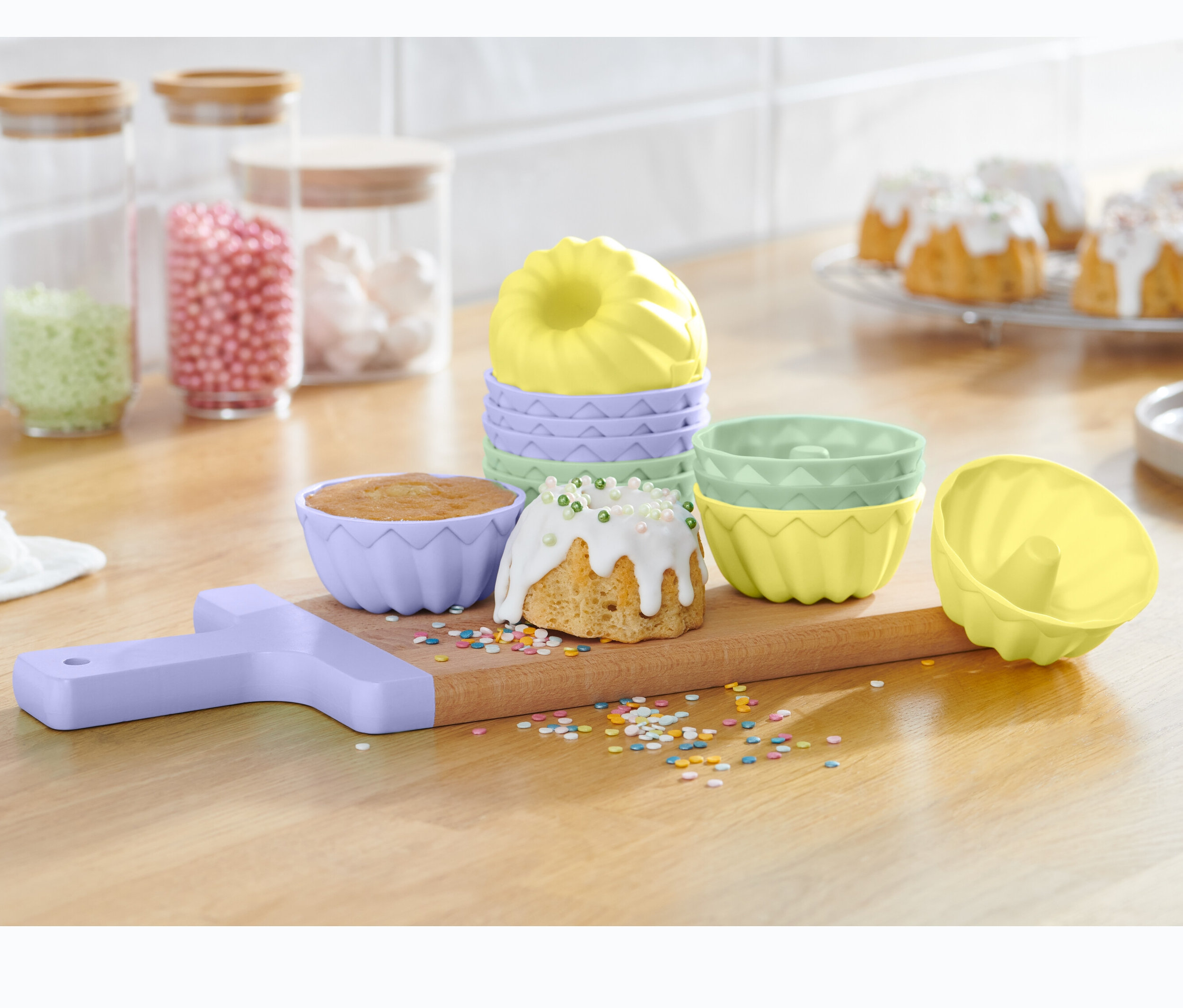 Egy fából készült deszkán 12 mini kuglóf sütőforma szettben látható, megsült kuglófokkal és szórással.