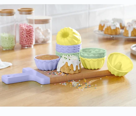 Egy fából készült deszkán 12 mini kuglóf sütőforma szettben látható, megsült kuglófokkal és szórással.